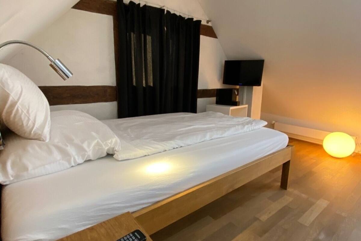 A bedroom with a bed, a bedside table with a remote control, a lamp, and a TV on a stand.