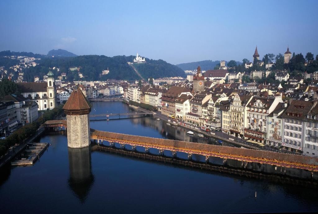 A river with a bridge and a tower, surrounded by historic buildings and a hill with a castle.