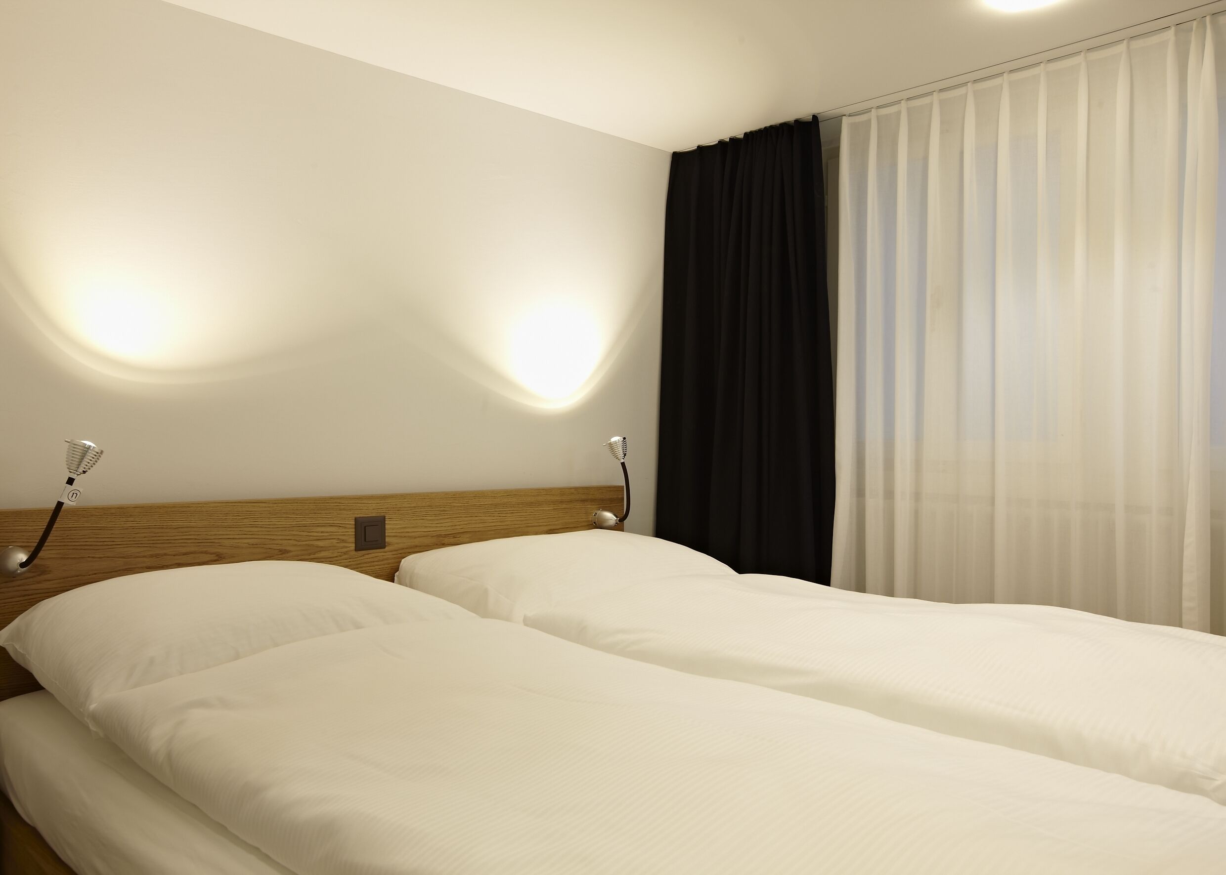 Camera d'albergo con due letti singoli, una testiera in legno, luci da lettura fissate al muro e un'ampia finestra con tende trasparenti.
