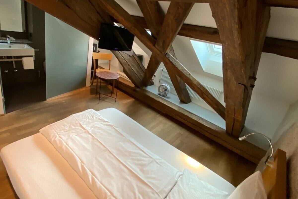 A loft bedroom with a wooden beam ceiling, a bed with white bedding, and a small table with a chair.