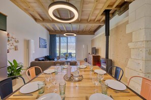 A dining area with a wooden table set for a meal, surrounded by chairs, with a view of the outdoors through large windows.