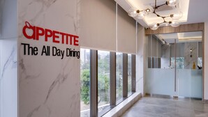 A modern hotel lobby with a marble wall, a sign reading "Cappetite The All Day Dining", and a large window offering a view of greenery.