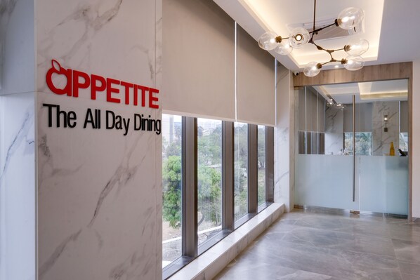 A modern hotel lobby with a marble wall, a sign reading "Cappetite The All Day Dining", and a large window offering a view of greenery.