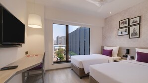 A hotel room with a TV, desk, two beds, a window with a city view, and framed pictures on the wall.