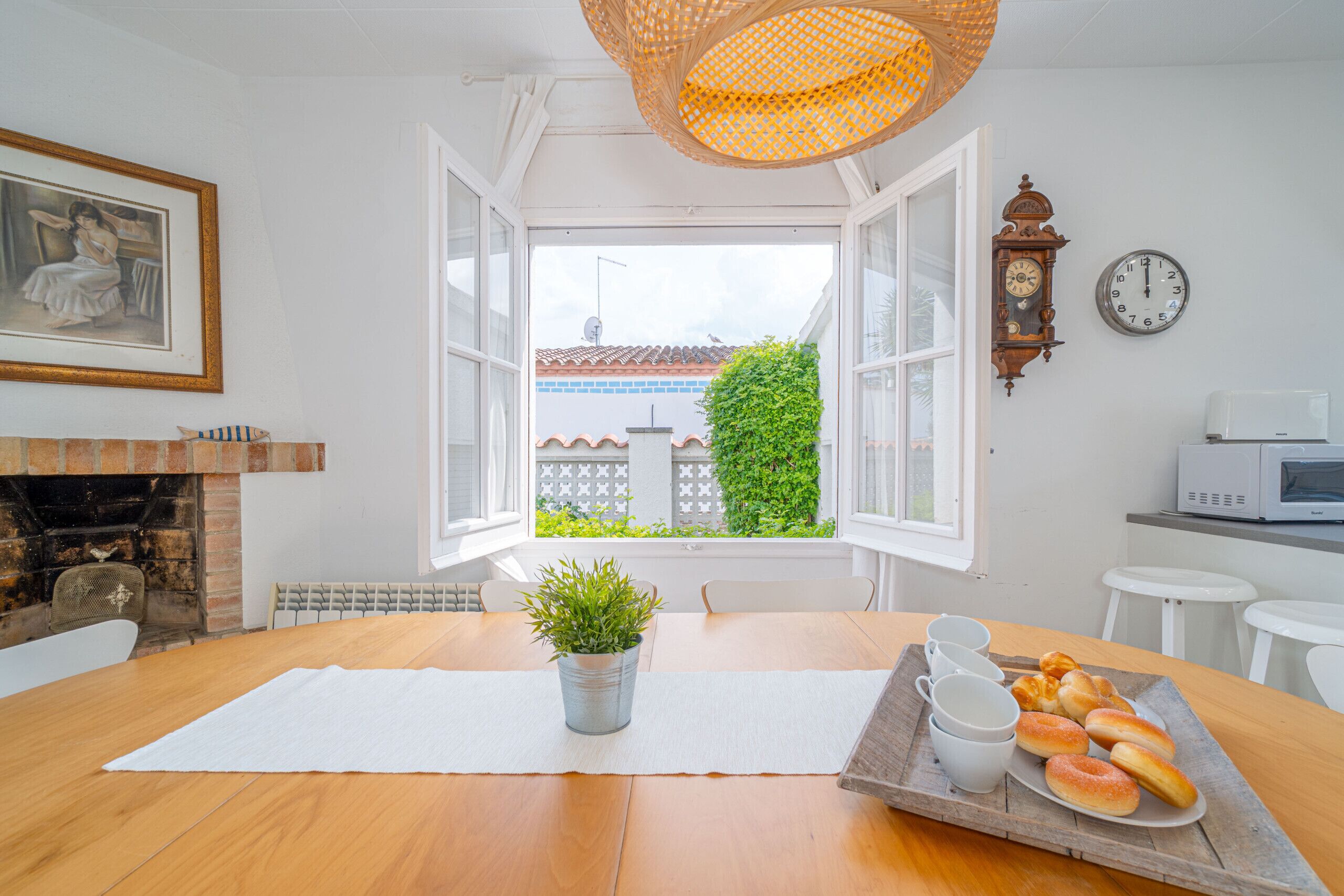 A dining area with a wooden table, a potted plant, a tray of bread, and a view of a building through open windows.