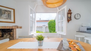 Un comedor con una mesa de madera, una planta en maceta, una bandeja de pan y vistas de un edificio a través de ventanas abiertas.