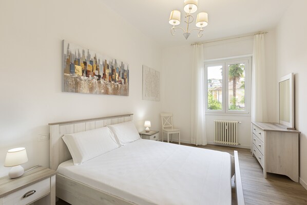 A bedroom with a bed, bedside table, lamp, chair, window, radiator, and a mirror. - Apartment Bononia (Stresa)