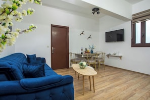 Un salon avec un canapé bleu, une table à manger en bois avec des chaises, un téléviseur fixé au mur et une porte.