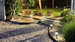 A well-lit garden pathway with a tree adorned with lights, surrounded by gravel and various shrubs. - COSY house 600 m from the beach, ideal for families - (Agon-Coutainville)