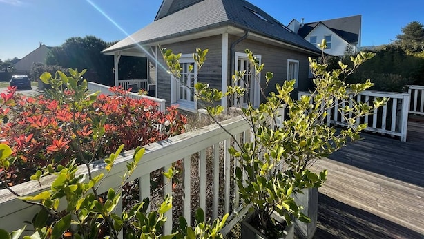 A two-story house with a grey roof and white trim, surrounded by a wooden deck and red flowering plants. - COSY house 600 m from the beach, ideal for families - (Agon-Coutainville)