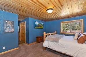 A bedroom with a wooden ceiling, a bed with white bedding, a small nightstand, and a window with blinds. - Situated in the Heart of Big Bear! Fully Fenced Yard! Pool Table! EV Charger! (Big Bear Lake)