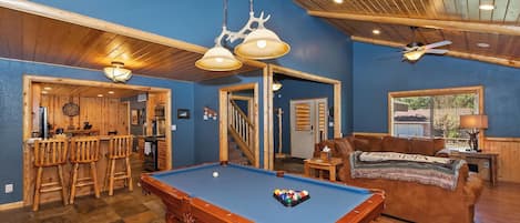 A pool table in a room with wooden flooring, a bar area, and a seating area with a couch.