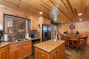 A kitchen with wooden cabinets, a stainless steel refrigerator, a sink, and a dining area with chairs and a table. - Situated in the Heart of Big Bear! Fully Fenced Yard! Pool Table! EV Charger! (Big Bear Lake)