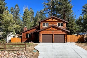 A two-story log cabin with a garage, surrounded by a wooden fence and trees. - Situated in the Heart of Big Bear! Fully Fenced Yard! Pool Table! EV Charger! (Big Bear Lake)