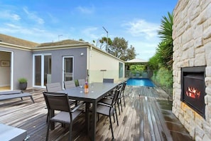 A wooden deck with a dining table and chairs, a swimming pool, and a stone wall with a fireplace.