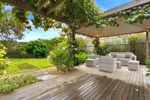 An outdoor patio with a wooden deck, white wicker furniture, and a pergola covered with vines. - Banksia House (Blairgowrie)