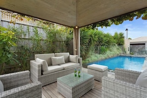An outdoor patio area with a wicker sofa, ottoman, and coffee table, a swimming pool, and lush greenery.