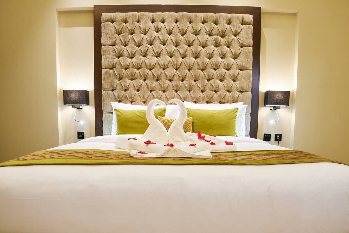Habitación de hotel con una cama grande, cabecera capitonada, almohadas verdes y una decoración de toallas con forma de cisne.