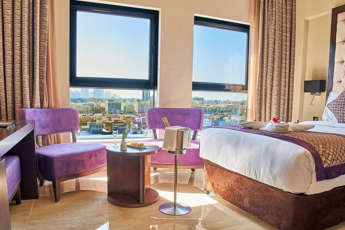 Habitación de hotel con una cama, dos sillones morados, una mesita redonda con una botella y copas, y vista a un paisaje urbano a través de amplias ventanas.