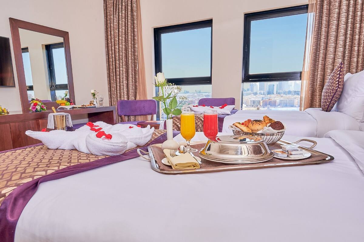 Habitación de hotel con una cama grande, una mesa de comedor con productos para el desayuno y vistas a la ciudad a través de las ventanas.