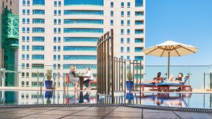 Een zwembad op het dak met ligbedden en parasols, omringd door een hoog gebouw met balkons.