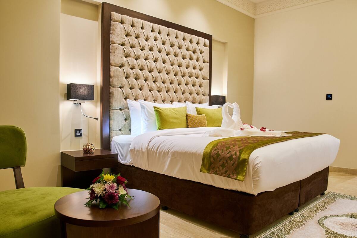 Habitación de hotel ordenada con una cama grande, una mesita de noche, una silla verde y una pequeña mesa redonda con un arreglo floral.