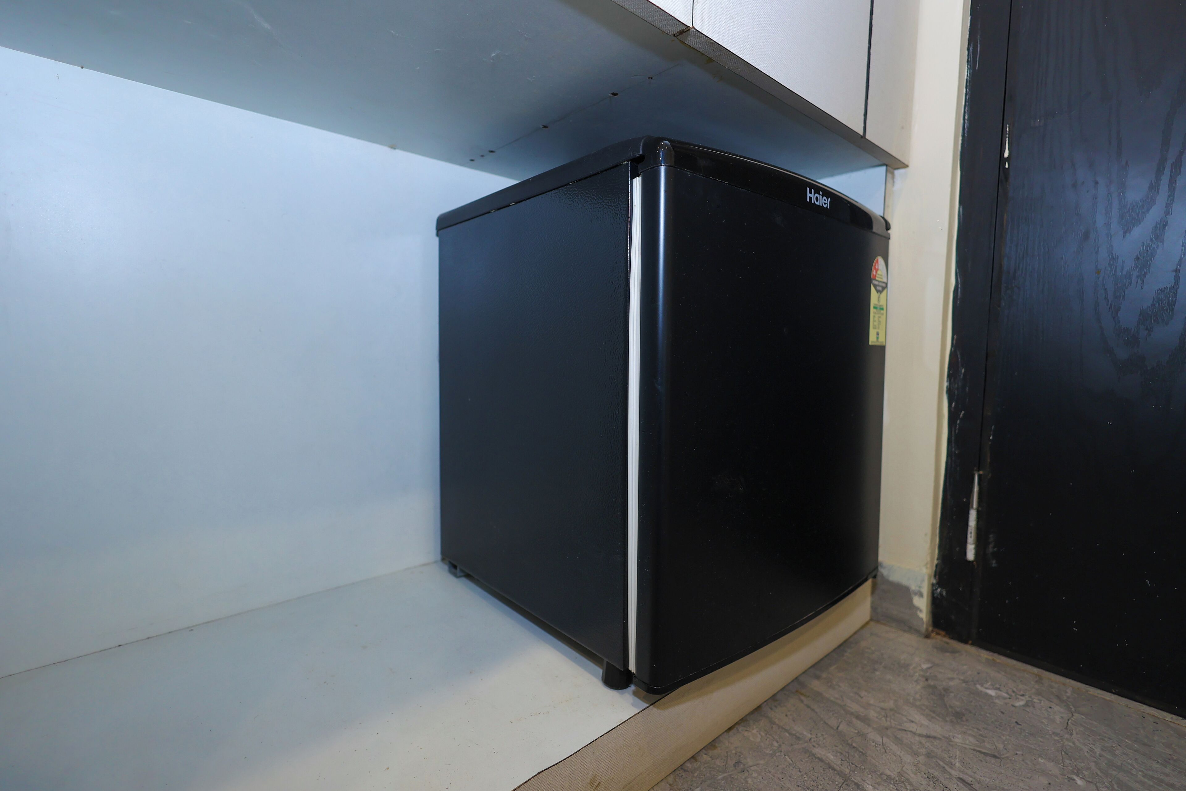 A black refrigerator in a corner of a room with white walls and a black door.