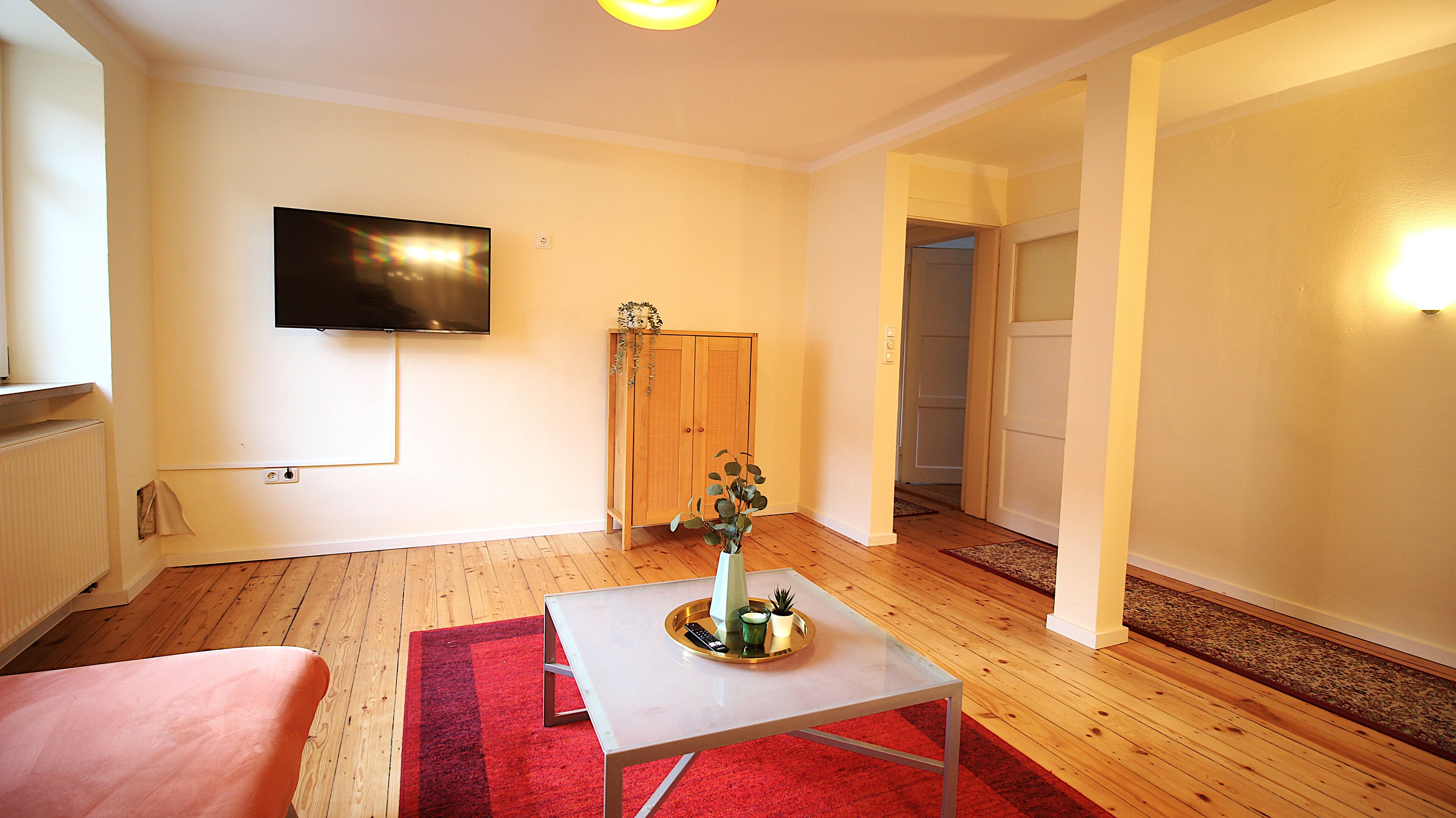 A living room with a flat-screen TV mounted on the wall, a wooden floor, a red and black rug, a small table with a vase and a tray, and a wooden wardrobe.