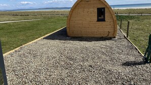 Una pequeña cabaña de madera con ventana, rodeada por un camino de grava y un campo cubierto de hierba, con vistas a una playa y montañas lejanas.