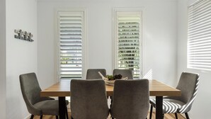 A dining area with a wooden table and four grey chairs, two windows with white blinds, and a small decorative piece on the wall.