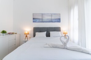 Een netjes opgemaakt bed met witte linnens, twee handdoeken in de vorm van zwanen, een grijs hoofdeinde, nachtkastjes met lampen, een kleine plant en een schilderij van een zeegezicht aan de muur.