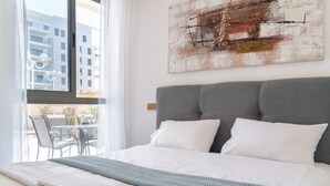 Une chambre d’hôtel moderne avec un grand lit, une tête de lit grise et une vue sur la ville grâce à une grande fenêtre.