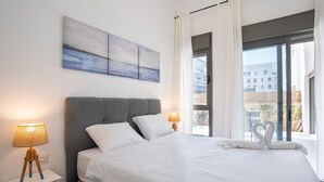 Une chambre d’hôtel moderne avec un grand lit, deux lampes de chevet, une table de chevet avec des tiroirs, une tête de lit rembourrée de couleur grise et une vue sur la ville à travers de grandes fenêtres.
