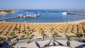Ein Strand mit vielen Reetsonnenstühlen und Sonnenschirmen, einer Pier mit Booten und einem klaren, blauen Meer.