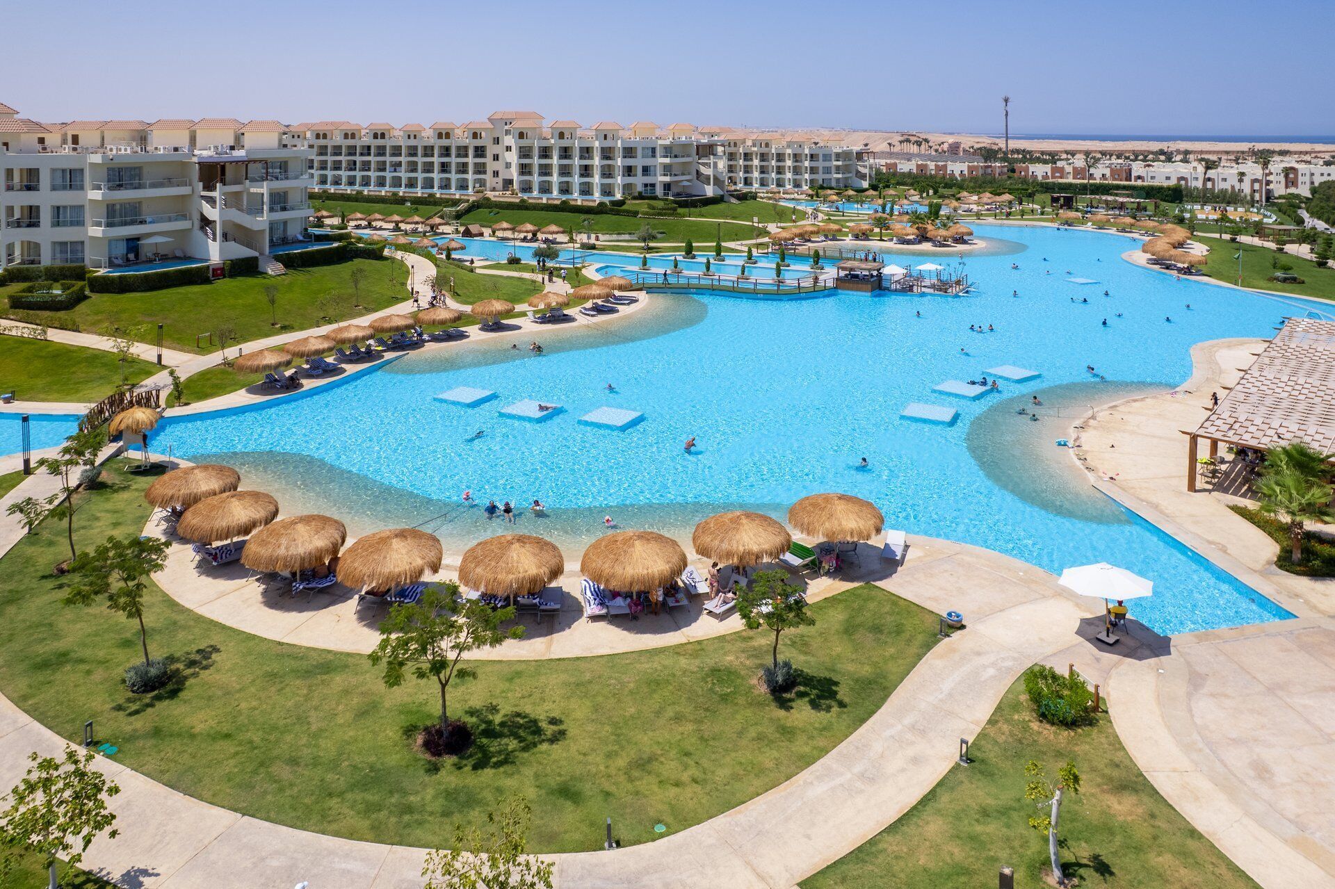 A large, multi-level hotel with a central swimming pool surrounded by sun loungers and thatched umbrellas.