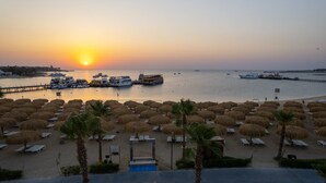 A beach with rows of sun loungers and thatched umbrellas, a pier with boats, and a sunset over the water. - Xanadu Makadi Bay - High Class All Inclusive (Makadi Bay)