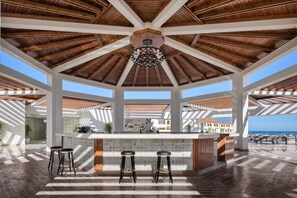 A spacious outdoor bar area with a high ceiling and wooden beams, featuring a central bar with stools and a view of the ocean.
