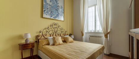 A bedroom with a bed, a nightstand, a lamp, a window with curtains, and a wall-mounted artwork.