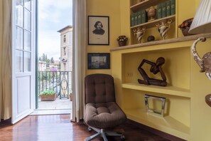 A room with a large window, a chair, a shelf with decorative items, and a framed picture on the wall.