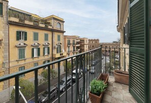 A balcony with a view of a street, potted plants, and a building with a yellow facade.
