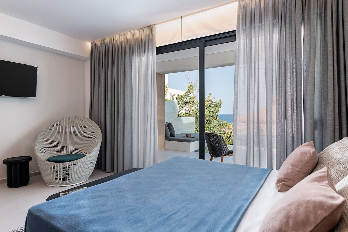 A modern bedroom with a bed, a wicker chair, a small table, and a view of the sea through large windows.