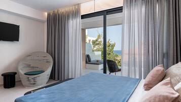 A modern bedroom with a bed, a wicker chair, a small table, and a view of the sea through large windows.