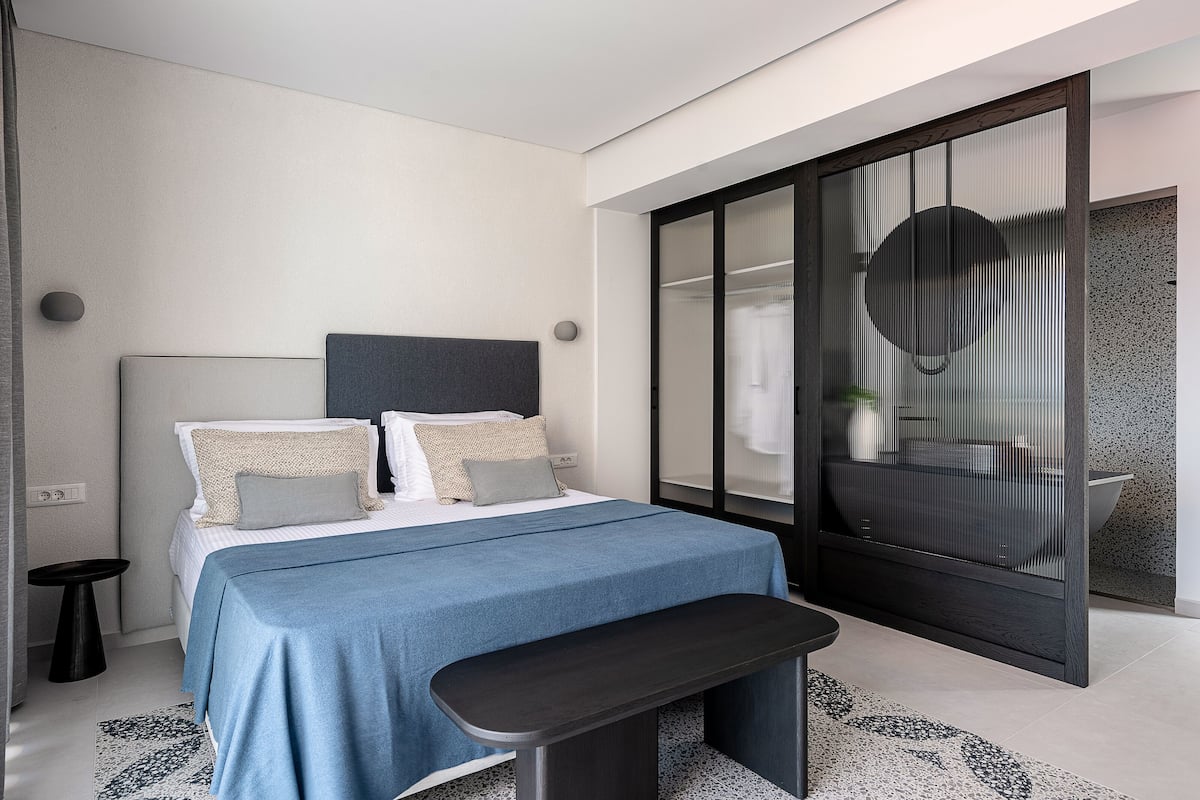 A modern bedroom with a large bed, a bench, a small side table, and a sliding glass door leading to a bathroom.