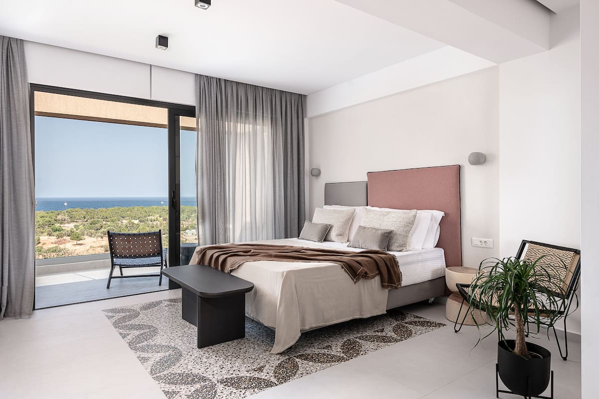 A modern bedroom with a large bed, a bench, a chair, a potted plant, and a view of the sea.