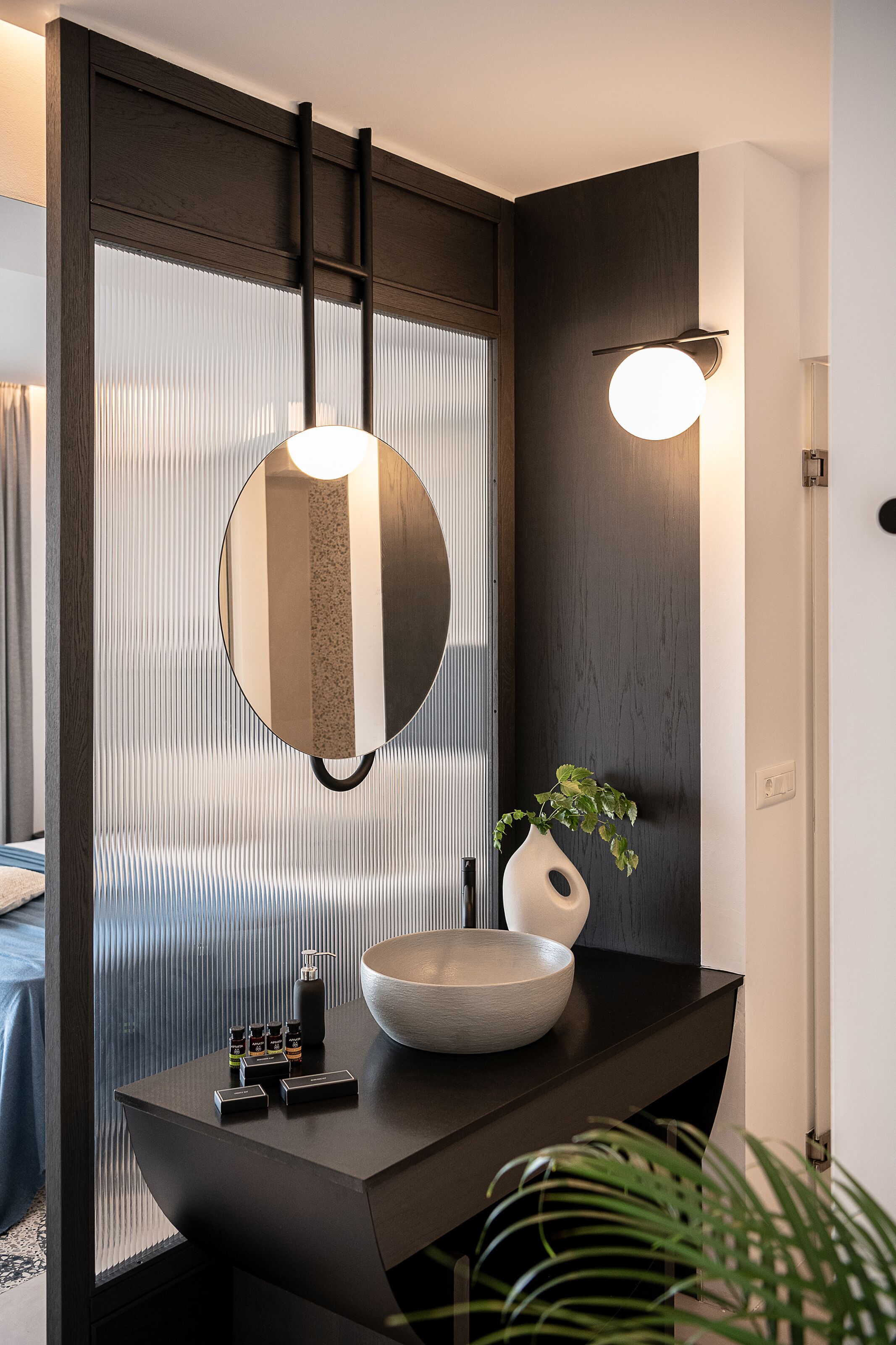 Un baño moderno con tocador de madera oscura, un espejo redondo, un lavabo tipo cuenco y una lámpara fijada a la pared.