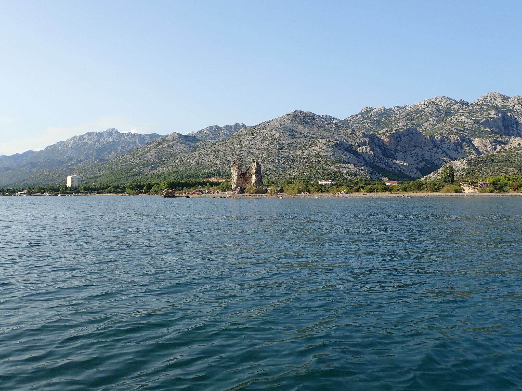 A coastal view with a rocky shoreline, a historic stone structure, and a mountainous backdrop.