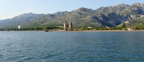 A coastal view with a rocky shoreline, a historic stone structure, and a mountainous backdrop.