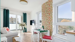 Una sala de estar moderna con sofá, mesa de comedor y sillas, televisor y ventanas amplias con cortinas.