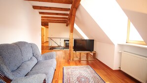 Un salon chaleureux avec un canapé, un téléviseur posé sur un meuble, des poutres en bois et une vue sur un escalier.