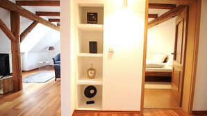 Une pièce avec des poutres en bois, un mur blanc, une étagère ornée d’objets décoratifs, un lit et une porte donnant sur une autre pièce.